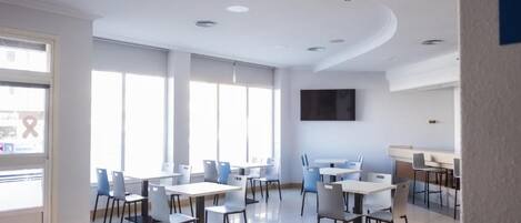 Un comedor moderno con mesas, sillas, ventanas amplias y un televisor de pantalla plana en la pared.