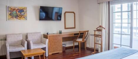 A hotel room with a bed, two armchairs, a desk, a TV, a window with curtains, and artwork on the wall.