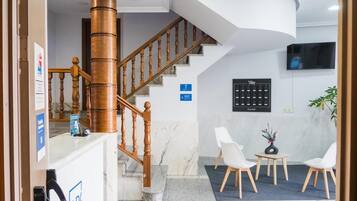 Zona de recepción con escalera de madera, mostrador de recepción, televisor montado en la pared y un área de estar con sillas y una mesa pequeña.