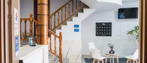 A reception area with a wooden staircase, a reception desk, a wall-mounted television, and a seating area with chairs and a small table.
