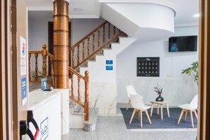 A reception area with a wooden staircase, a reception desk, a wall-mounted television, and a seating area with chairs and a small table. - Alda Arteixo (Abegondo)