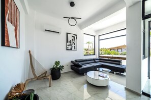 A spacious living room with a black sectional sofa, a round coffee table, and a hammock. - Elion Seaside Resort (Chalcis)