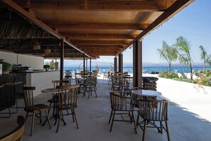 An outdoor patio with wooden tables and chairs, a bar area, and a view of the ocean. - Elion Seaside Resort (Chalcis)