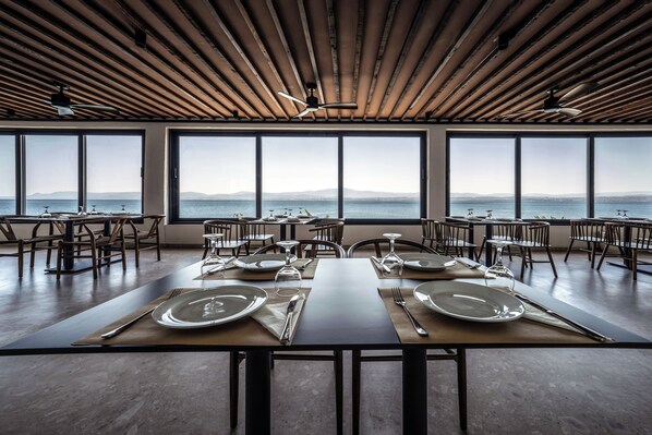 A dining area with tables set for four, large windows offering a view of the sea, and a wooden ceiling. - Elion Seaside Resort (Chalcis)