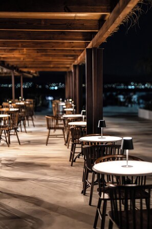 An outdoor patio area with round tables and chairs, wooden beams overhead, and a view of the night sky. - Elion Seaside Resort (Chalcis)