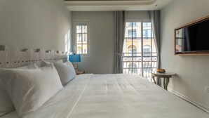 Habitación de hotel con una cama grande, un televisor montado en la pared, una mesita con una lámpara y una ventana con vistas a edificios.