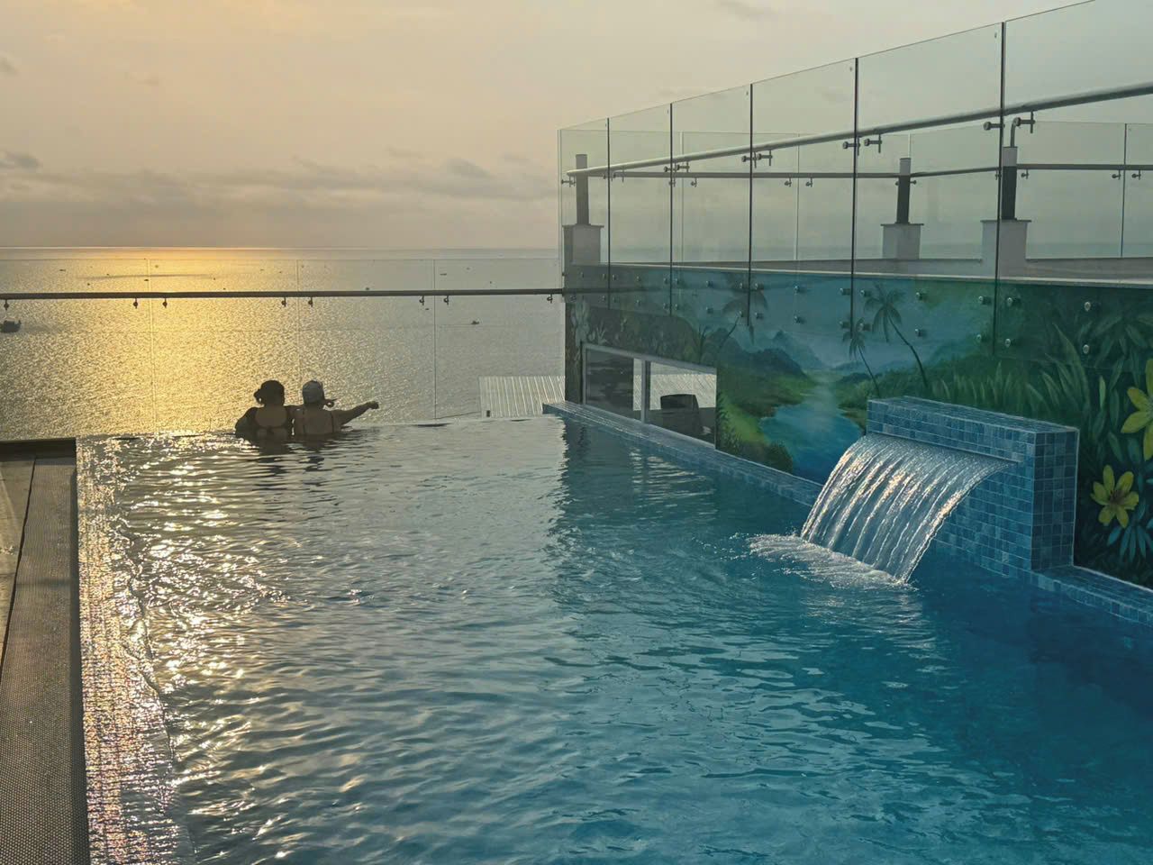 A swimming pool with a waterfall feature, a glass railing, and a view of the sea.