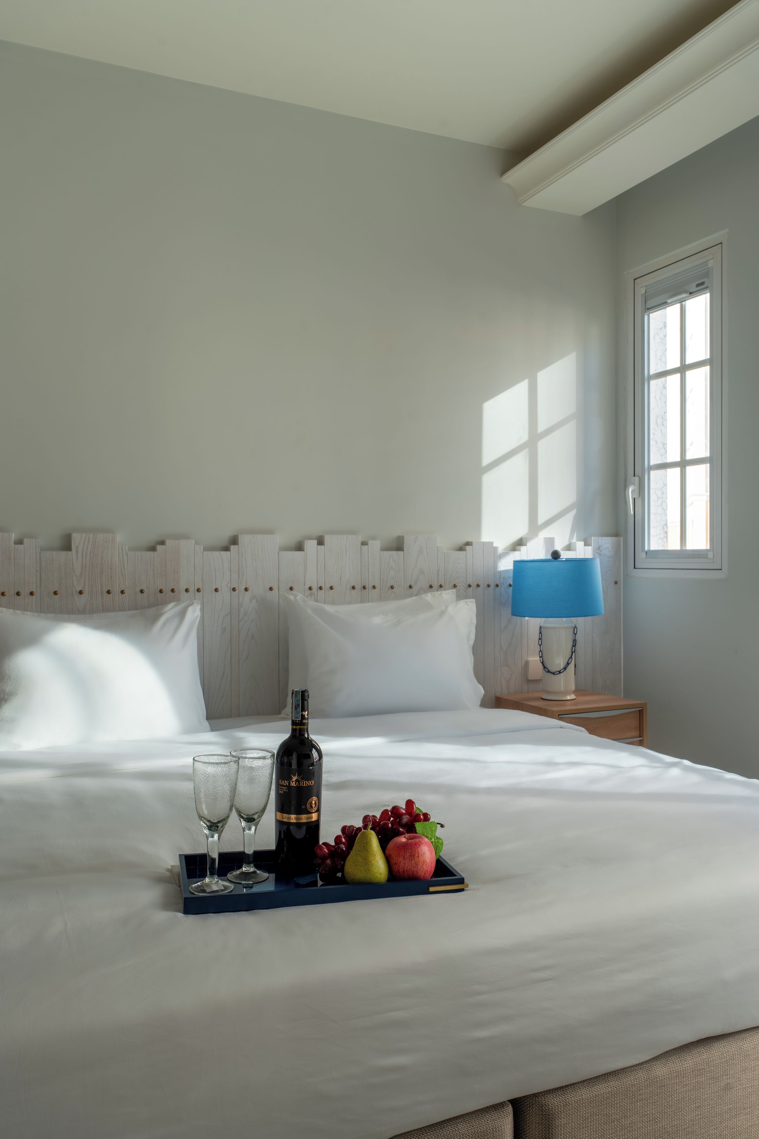 Un lit bien fait, avec un plateau de fruits et une bouteille, une lampe de chevet et une fenêtre laissant entrer la lumière du jour.