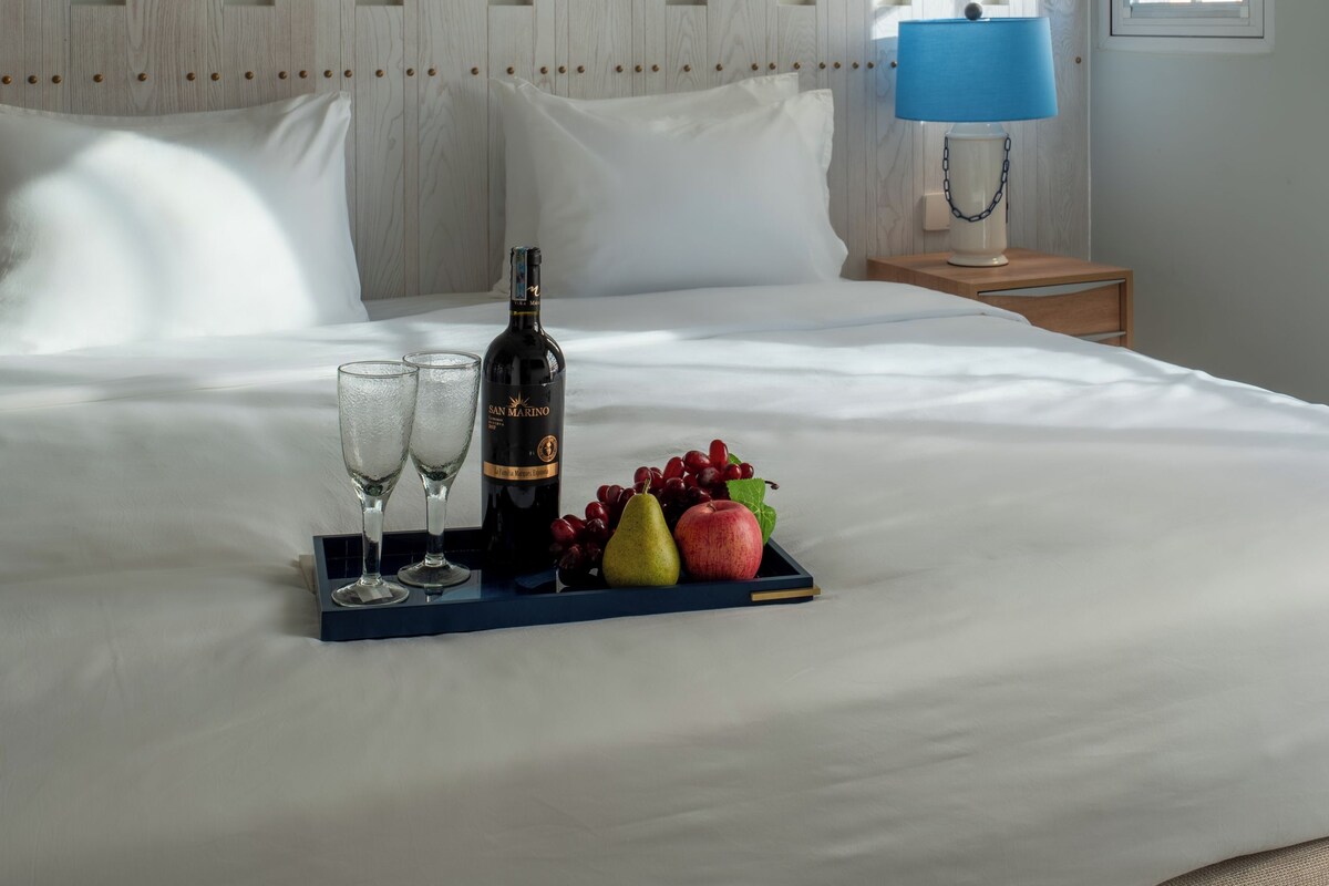 A neatly made bed with a tray of fruit and a bottle, a bedside lamp, and a window with daylight.