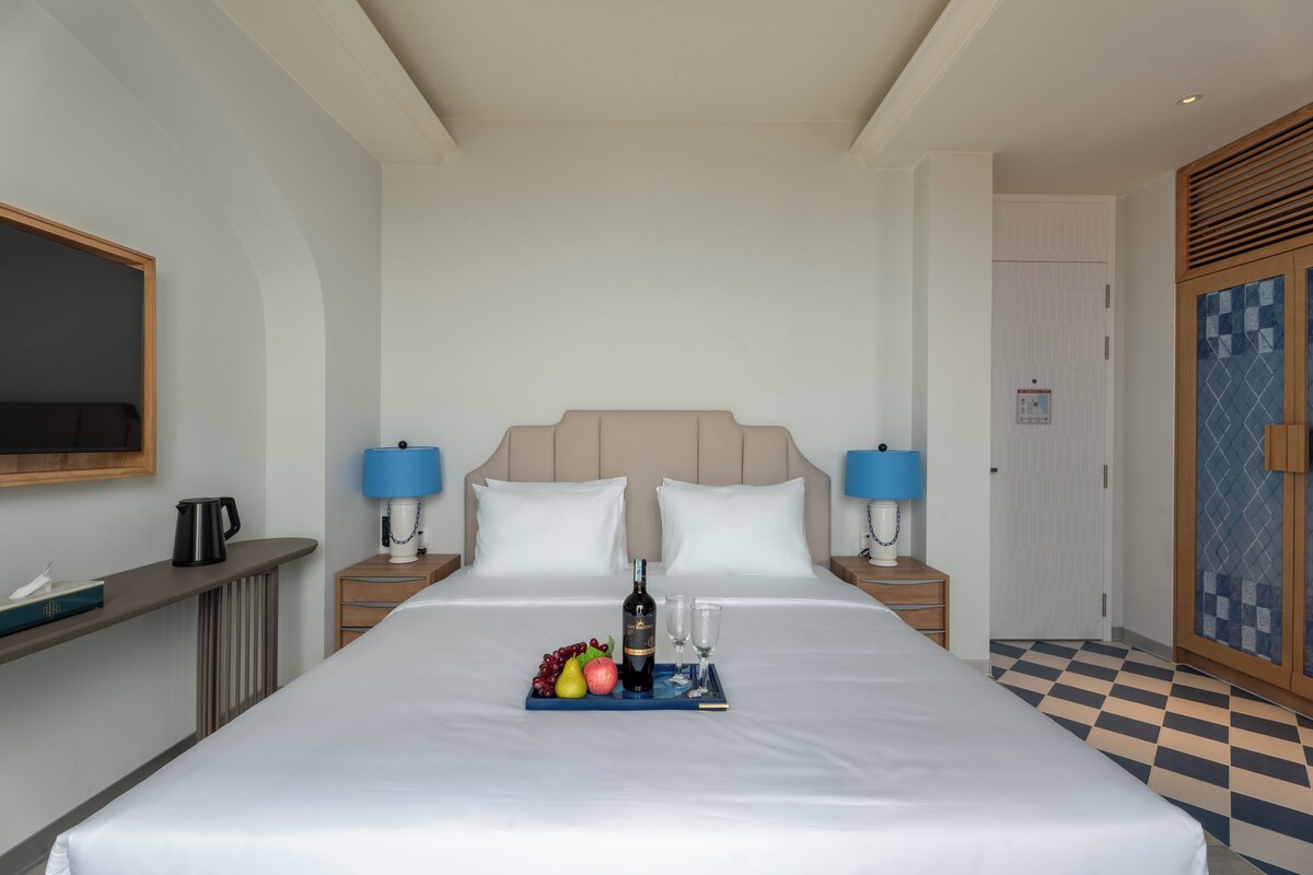 A hotel room with a large bed, two bedside tables with lamps, a minibar, and a check-in desk.
