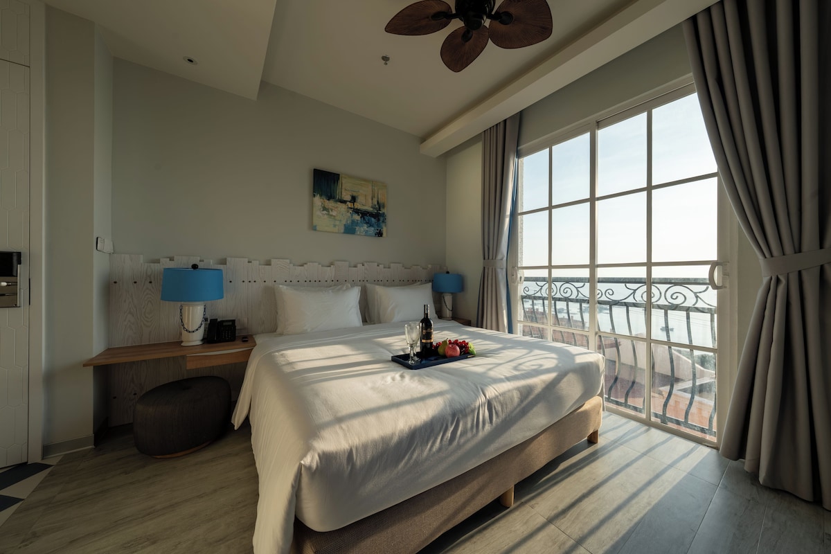 A hotel room with a large bed, bedside lamps, a painting, and a balcony with a view.