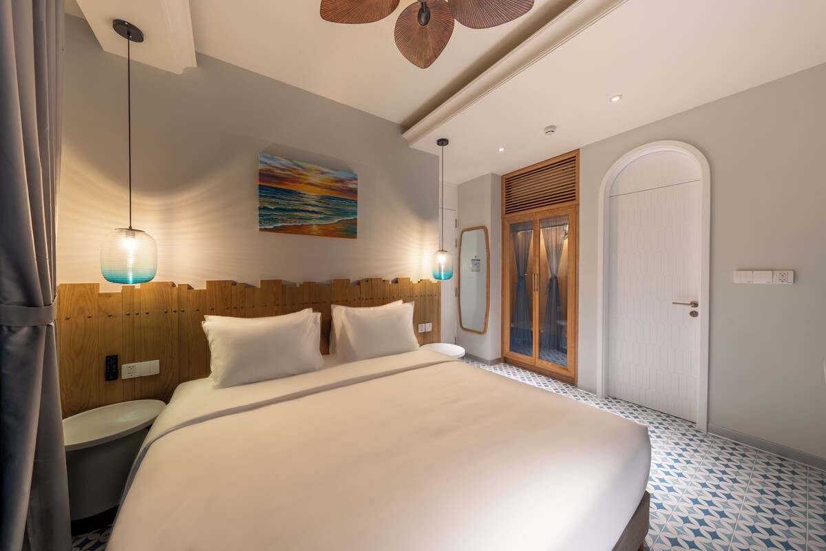 A modern hotel room with a large bed, wooden headboard, two wall-mounted lights, a painting, a mirror, and a door.