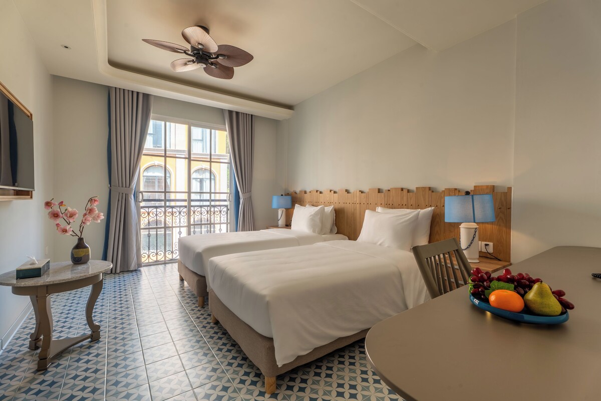 A hotel room with two beds, a dining table with fruit, a small table with a vase, and a balcony with curtains.