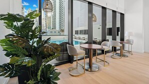 Ein modernes Hotelzimmer mit großen Fenstern, Blick auf einen Pool und einer Sitzecke mit runden Tischen und Stühlen.
