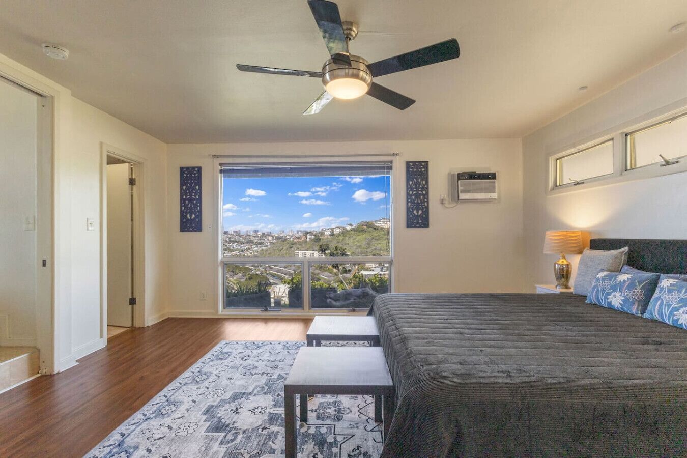 A bedroom with a bed, a ceiling fan, a window with a city view, and a small table.