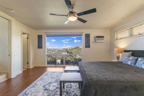 Ein Schlafzimmer mit Bett, Deckenventilator, Fenster mit Stadtblick und einem kleinen Tisch.