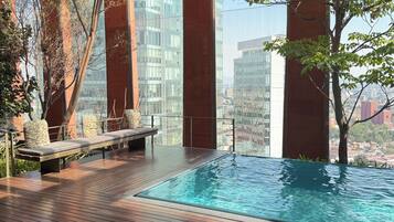 A rooftop infinity pool with wooden decking and seating, surrounded by glass walls and trees.