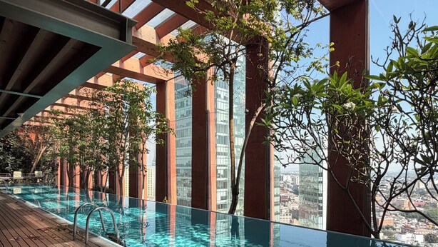 Une piscine à débordement située sur le toit, avec une terrasse en bois, entourée de murs en verre et offrant une vue sur le paysage urbain.
