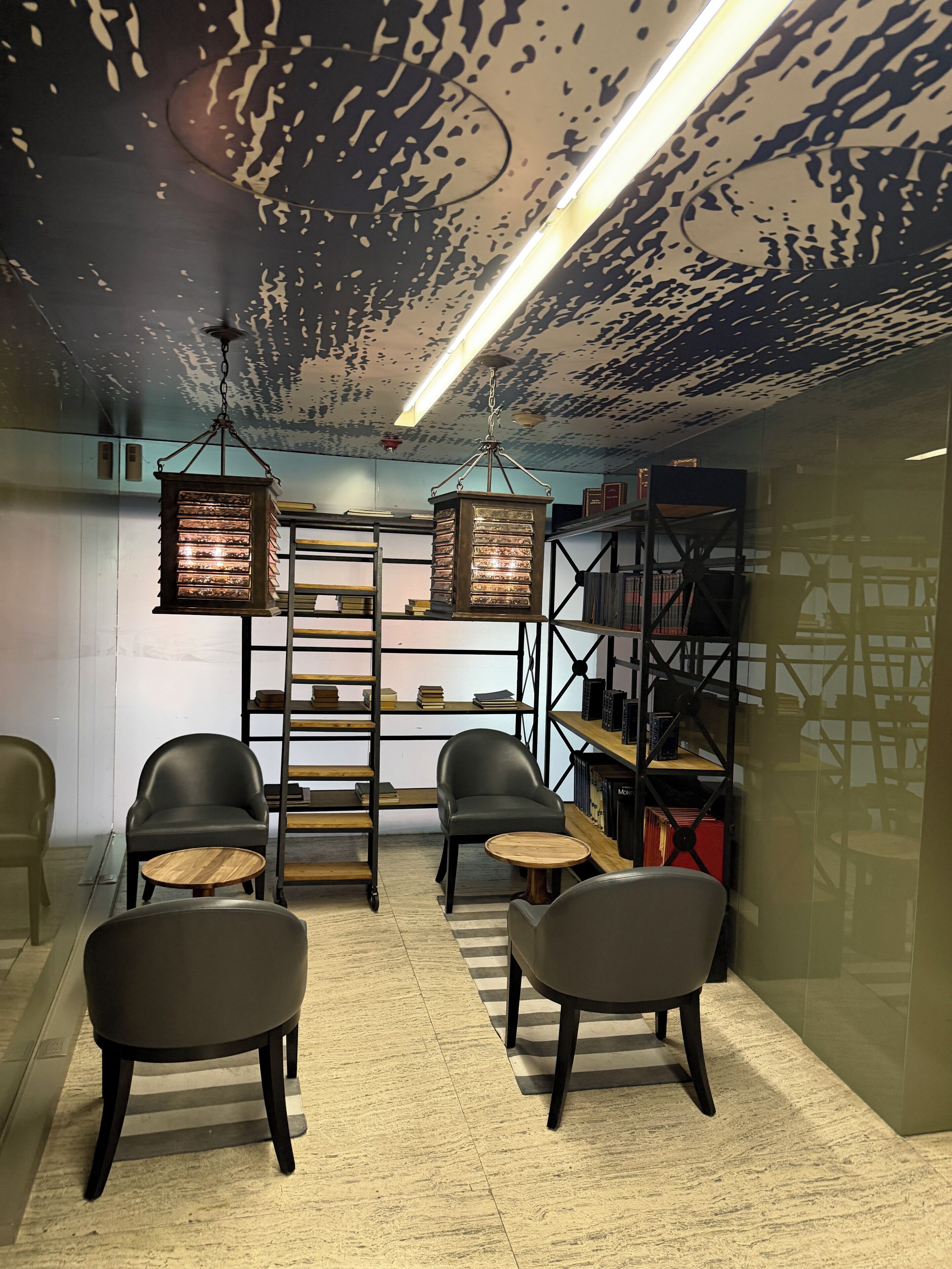 A modern interior with a ceiling with a unique pattern, two hanging lantern-style lights, a central wooden ladder, and a bookshelf with books and decorative items.