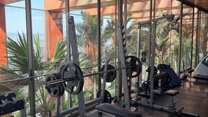 Une salle de sport équipée de matériel de musculation, de bancs et offrant une vue sur les palmiers et les immeubles grâce à de grandes fenêtres.