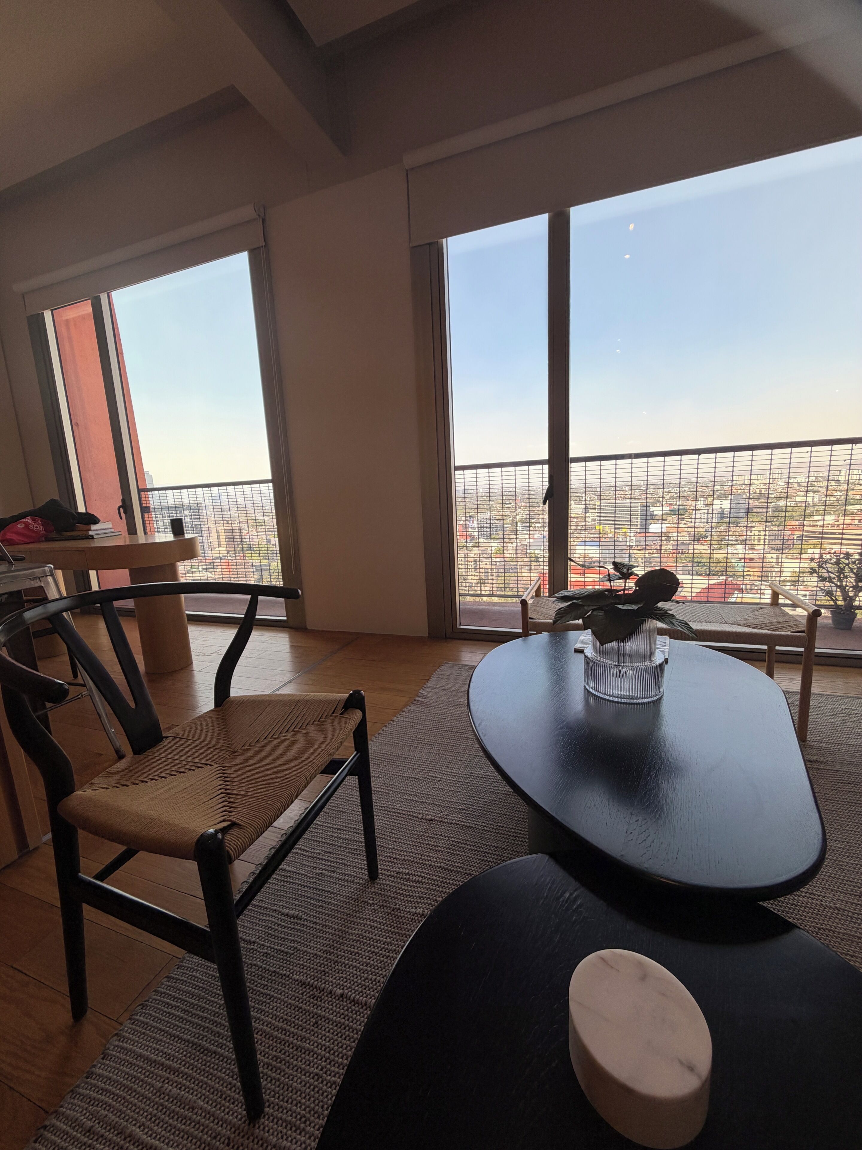 A modern living room with a dining table, chairs, a sofa, and large windows offering a city view.