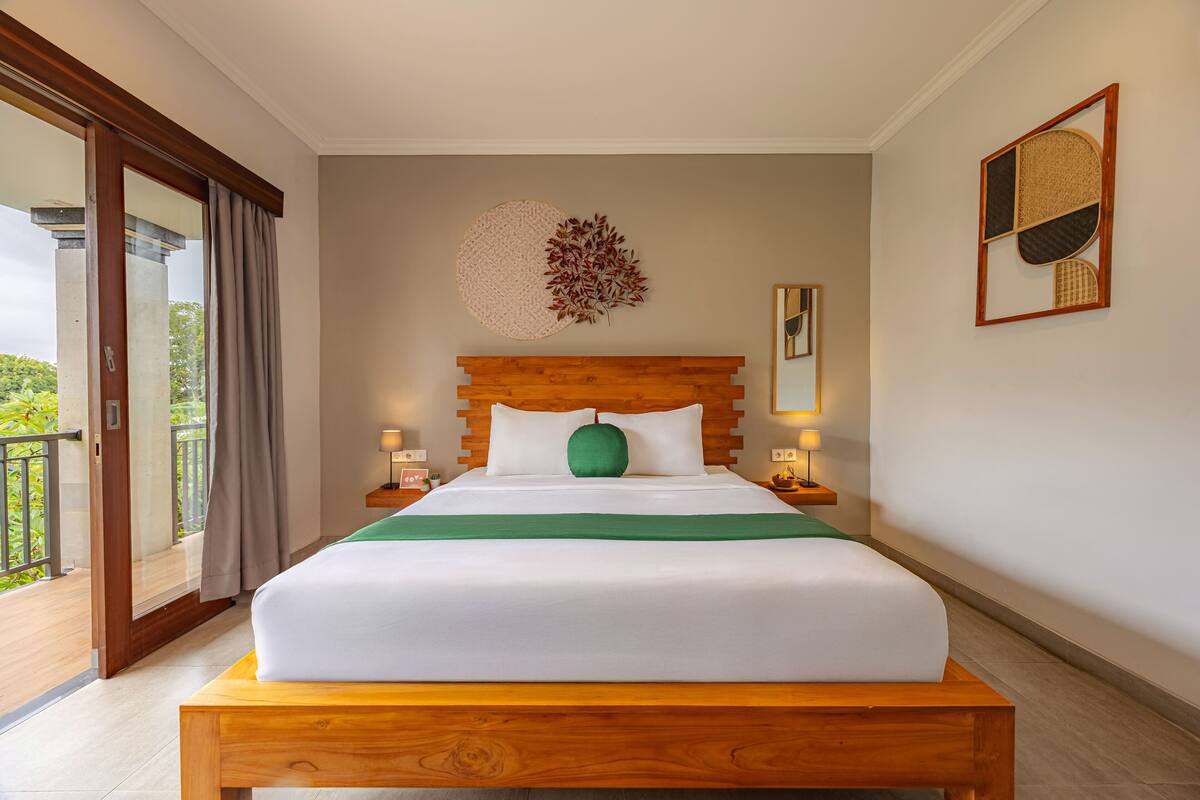 Un lit bien fait, avec une tête de lit en bois, un oreiller vert et un couvre-lit blanc. On y trouve deux tables de chevet avec des lampes, une œuvre d’art accrochée au mur au-dessus du lit, et une porte-fenêtre donnant sur un balcon, ornée de rideaux.