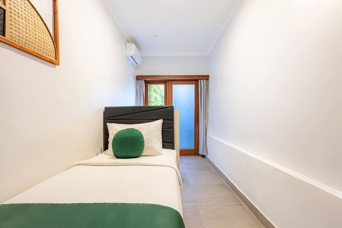 Un couloir étroit d’hôtel, avec un lit agrémenté d’un oreiller vert, un ventilateur de plafond et une fenêtre avec des rideaux.