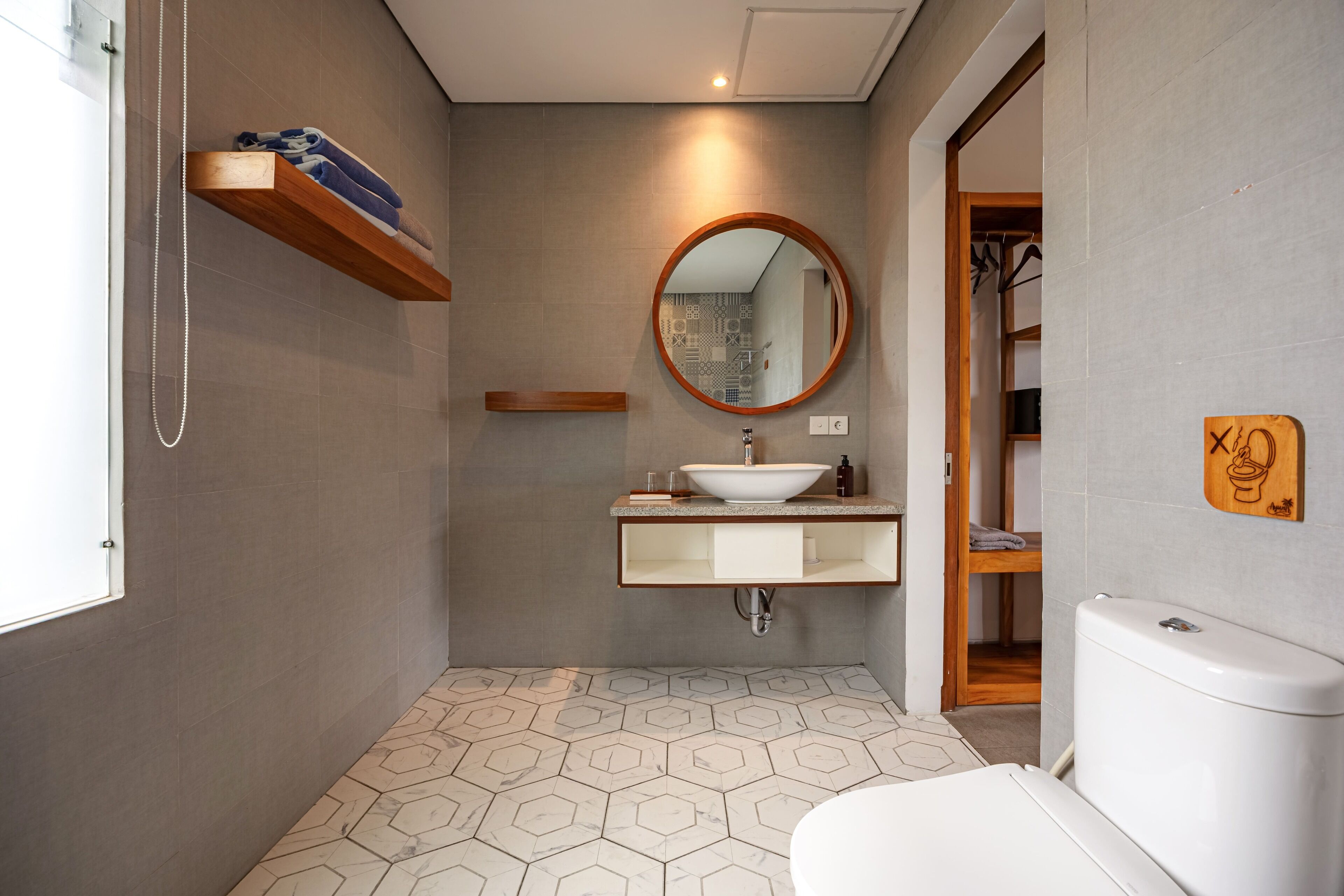 A modern bathroom with a hexagonal tile floor, a wooden shelf with towels, a round mirror, a white sink, and a toilet.
