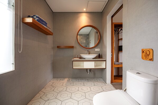 Un baño moderno con piso de baldosas hexagonales, un estante de madera con toallas, un espejo redondo, un lavamanos blanco y un inodoro.