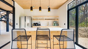 Eine moderne Küche mit Holzbalkendecke, drei Pendelleuchten, einem Kühlschrank und einer Kaffeestation.
