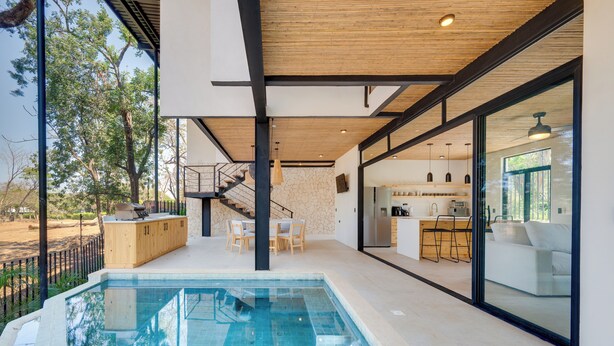 Ein moderner Innenpoolbereich mit Holzbalkendecke, eine Küche mit Bar und ein Essbereich mit einer Treppe, die zu einer erhöhten Ebene führt.