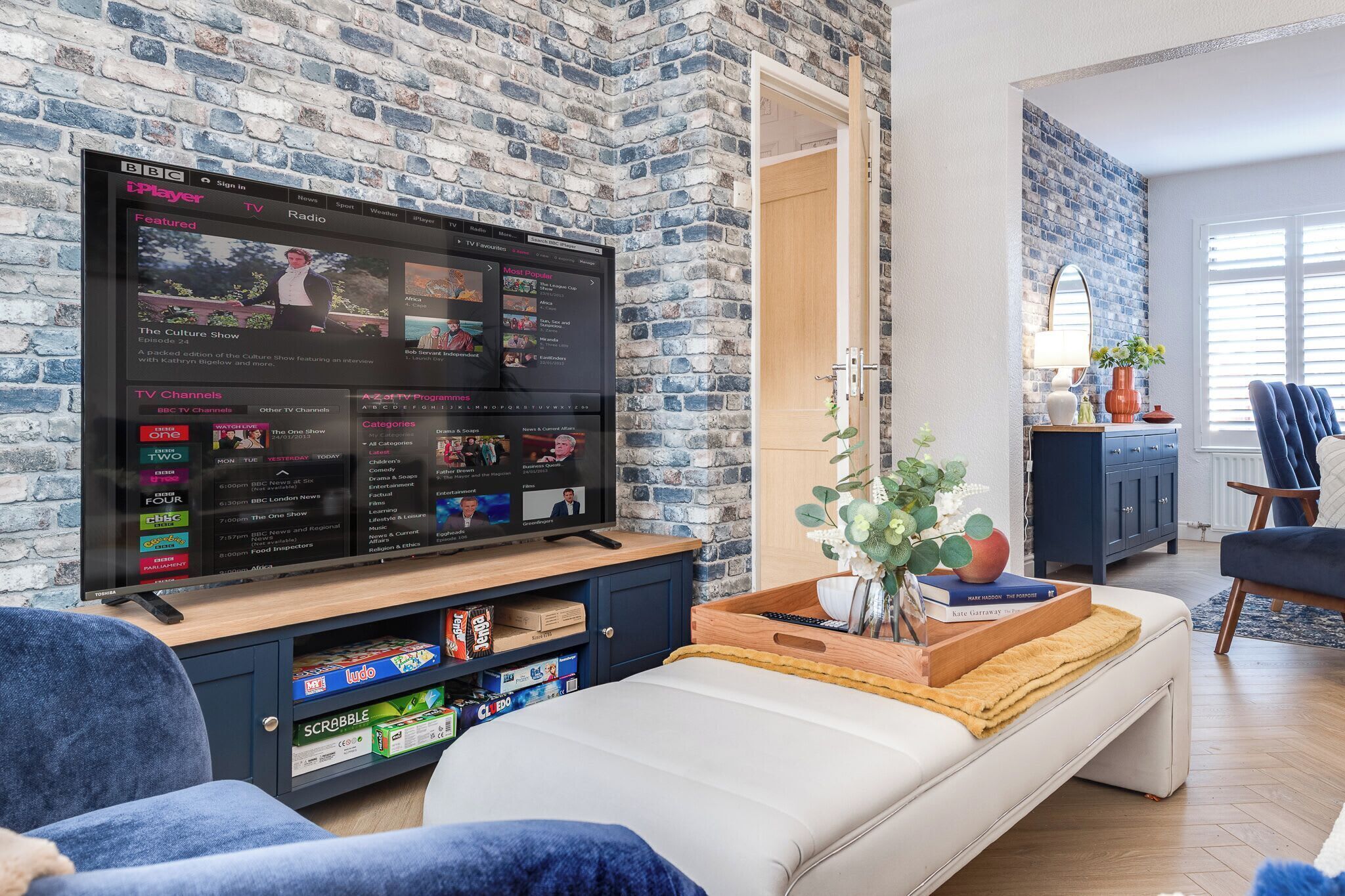 Ein Wohnzimmer mit einem großen Flachbildfernseher an einer Ziegelwand, einem Sofa, einem Couchtisch mit Büchern und einer Pflanze sowie einem blauen Sessel.