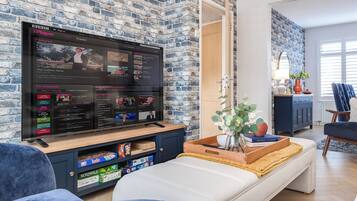 Ein Wohnzimmer mit einem großen Flachbildfernseher an einer Ziegelwand, einem Sofa, einem Couchtisch mit Büchern und einer Pflanze sowie einem blauen Sessel.