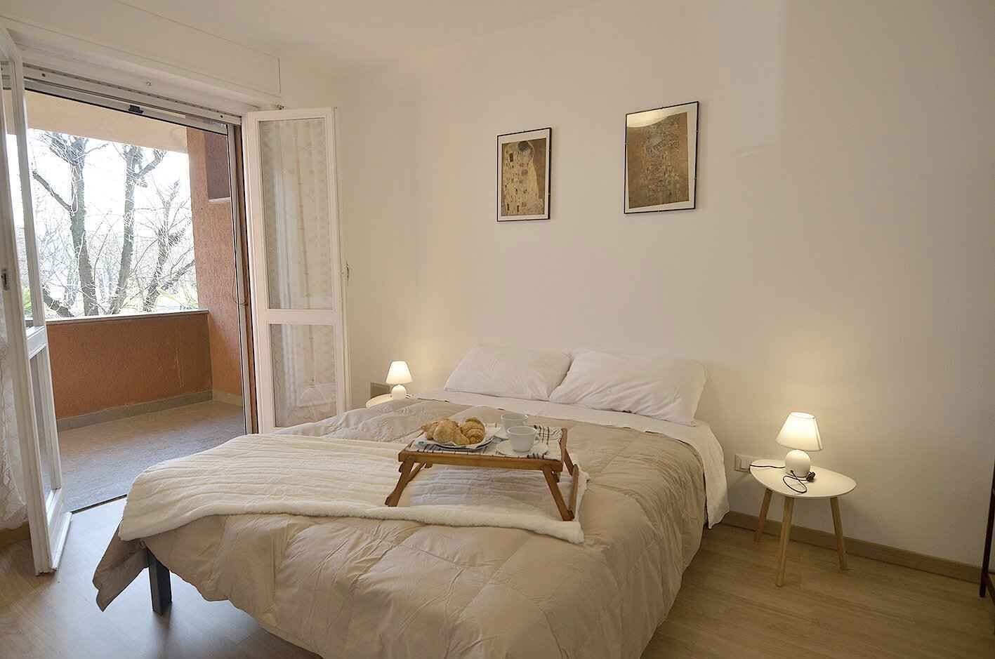 A bedroom with a bed, a bedside table with a lamp, a window with a view of trees, and two framed pictures on the wall.