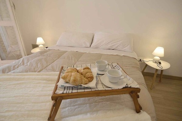 A neatly made bed with a tray of croissants and cups of coffee, a bedside table with a lamp, and a window with curtains.