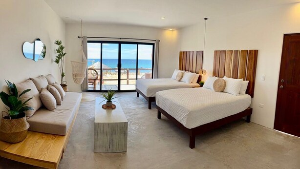 Habitació d'hotel amb un gran finestral corredís que dona a un balcó amb vista al mar. Hi ha dos llits amb roba de llit blanca, un sofà i una taula de fusta petita amb una planta.