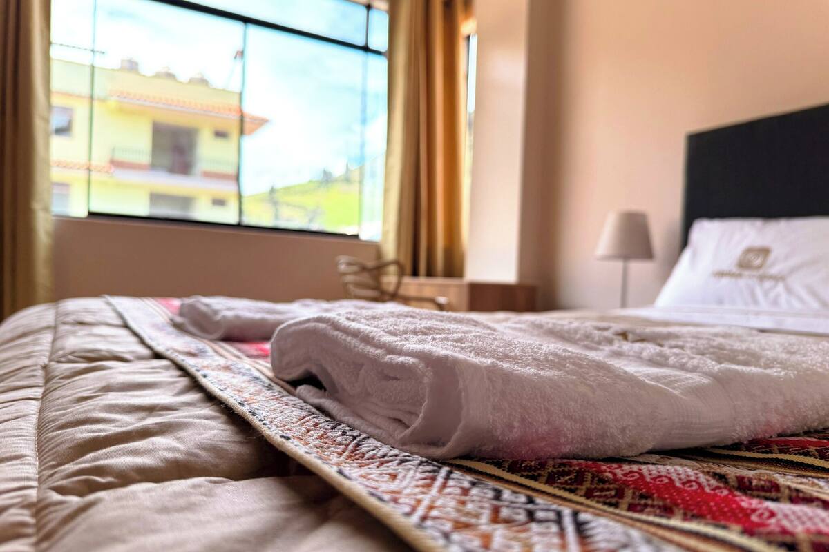 Una cama bien hecha con una colcha estampada, una ventana con vistas a edificios y una lámpara de noche.