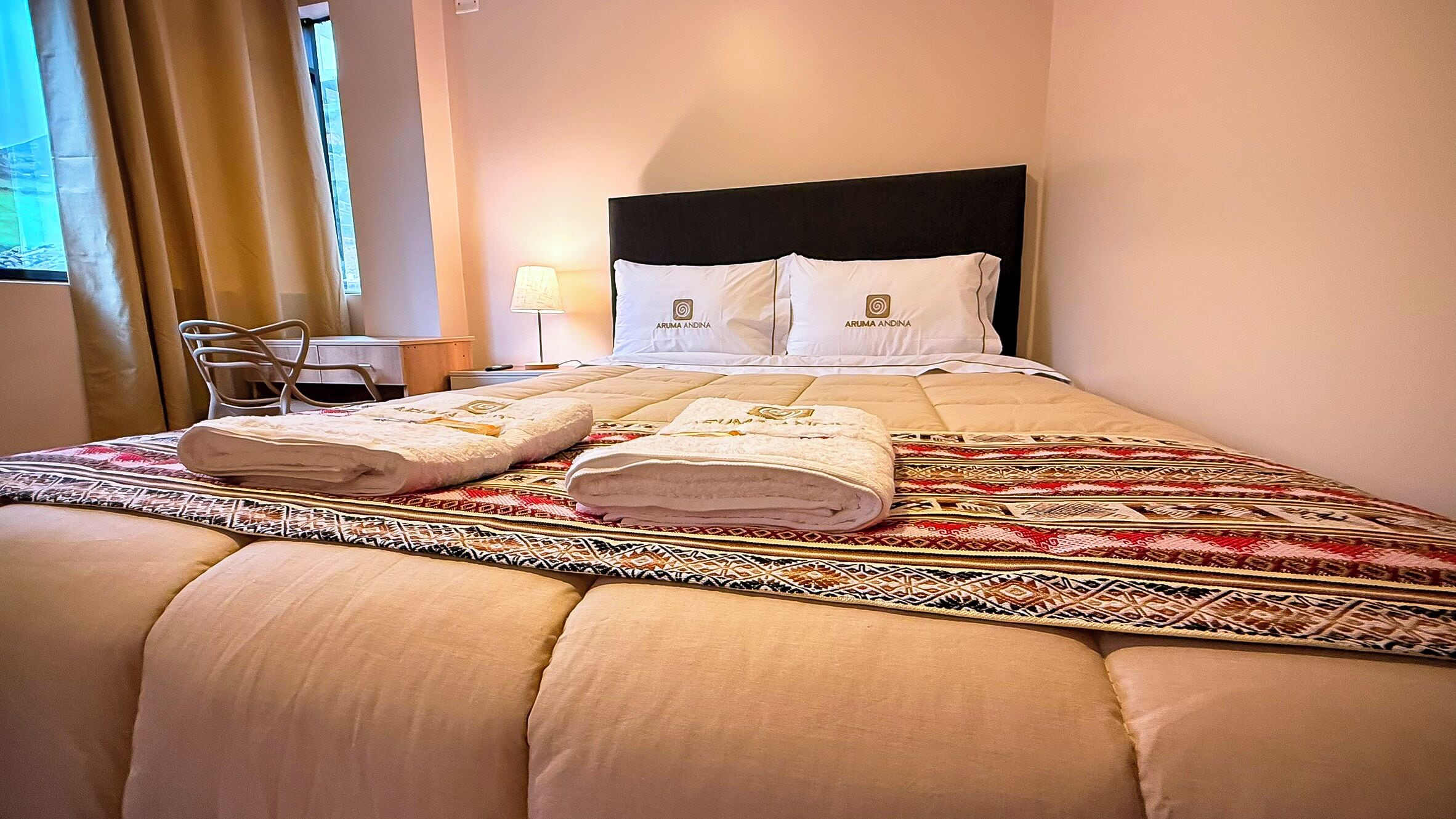 Cama bem arrumada com duas toalhas, cabeceira e mesa de cabeceira com abajur.