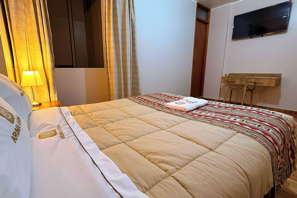 Habitación de hotel con cama, mesita de noche con lámpara, televisor montado en la pared y una puerta.
