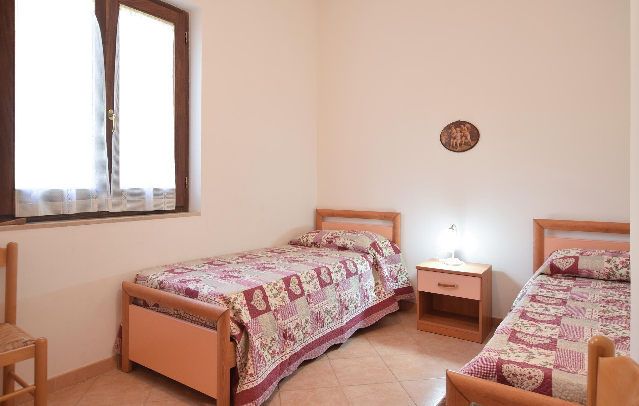 A bedroom with two wooden beds, a nightstand with a lamp, a window with blinds, and a wall-mounted picture.