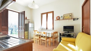 Una cocina compacta con refrigerador, una mesa de comedor con sillas, un sofá amarillo, un televisor y una ventana con cortinas.