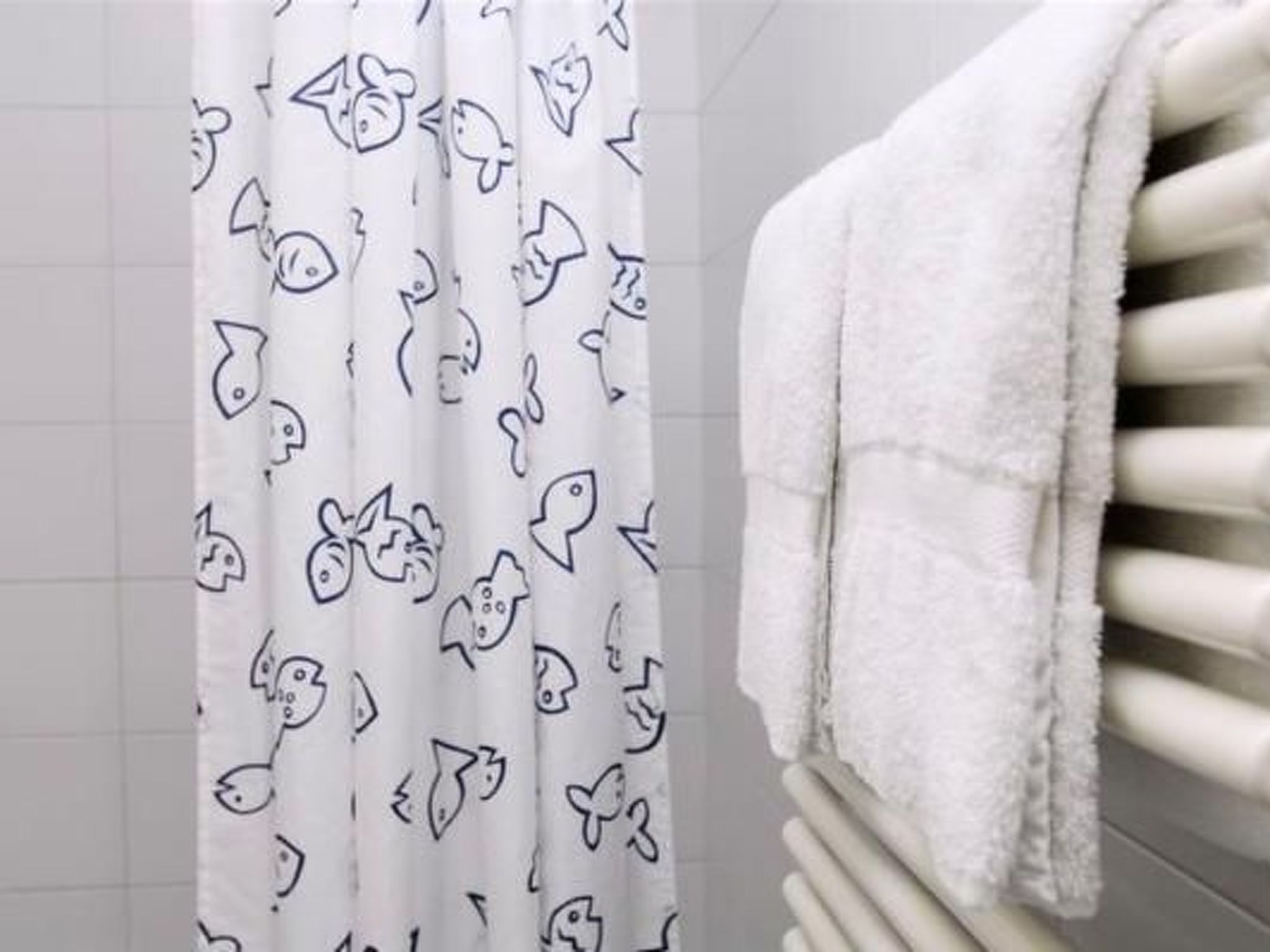 A bathroom with a white tiled wall, a white shower curtain with cartoon fish patterns, and a stack of white towels.