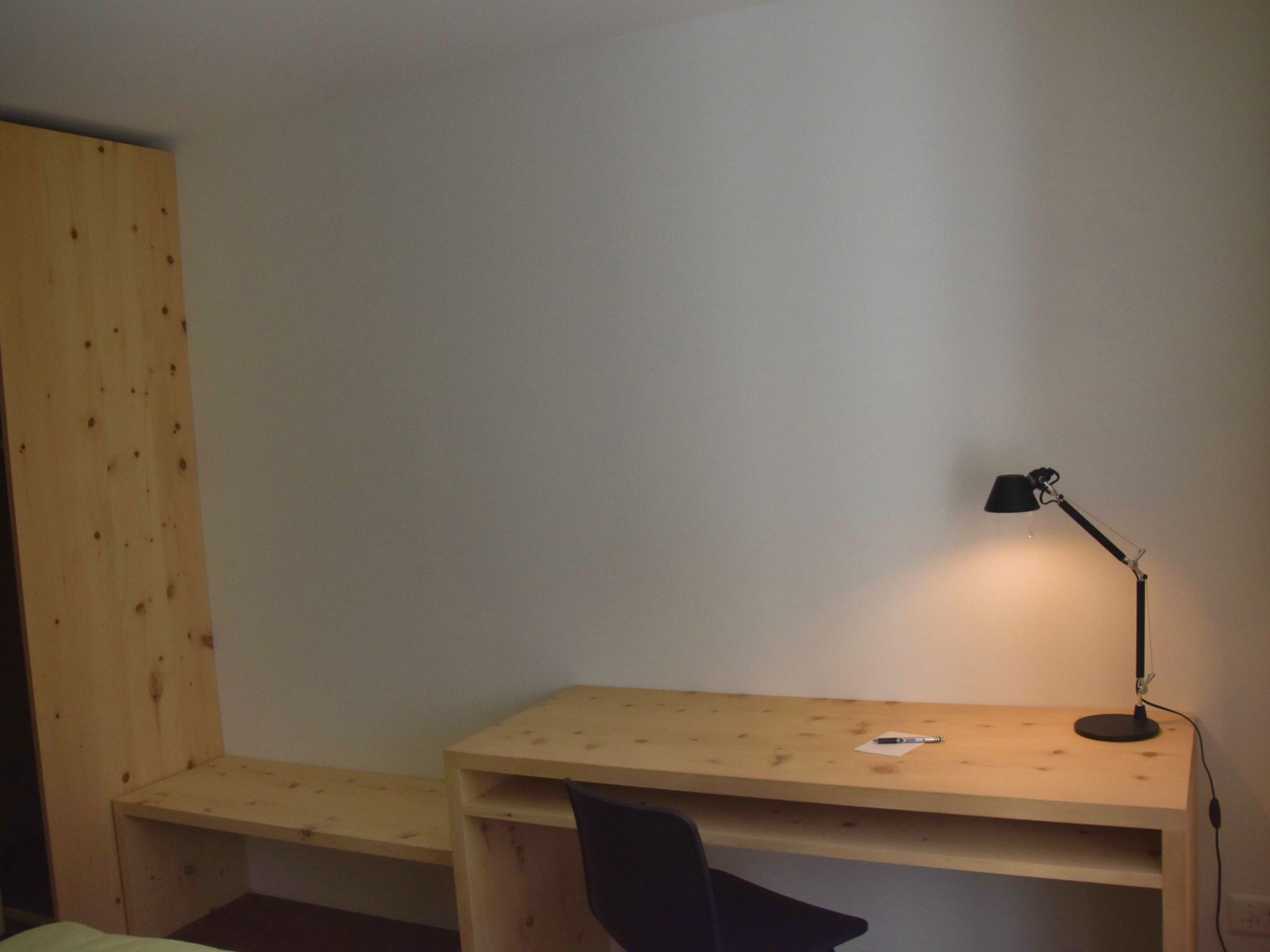 Une petite pièce simple, avec un bureau et une chaise en bois, une lampe de bureau noire et une cloison en bois.