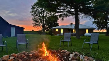 A campfire with chairs around it, a building with a metal roof, and trees in the background.