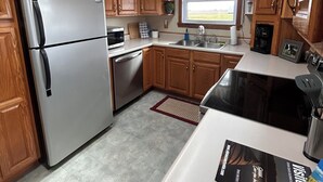 A kitchen with wooden cabinets, a stainless steel refrigerator, a dishwasher, a window with a view, and a countertop with a magazine.