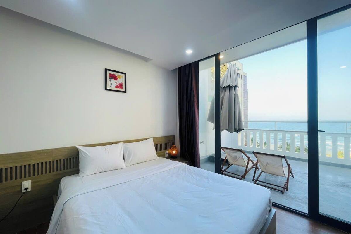 A hotel room with a large bed, a bedside table with a lamp, a window with a view of the sea, and a balcony with chairs.