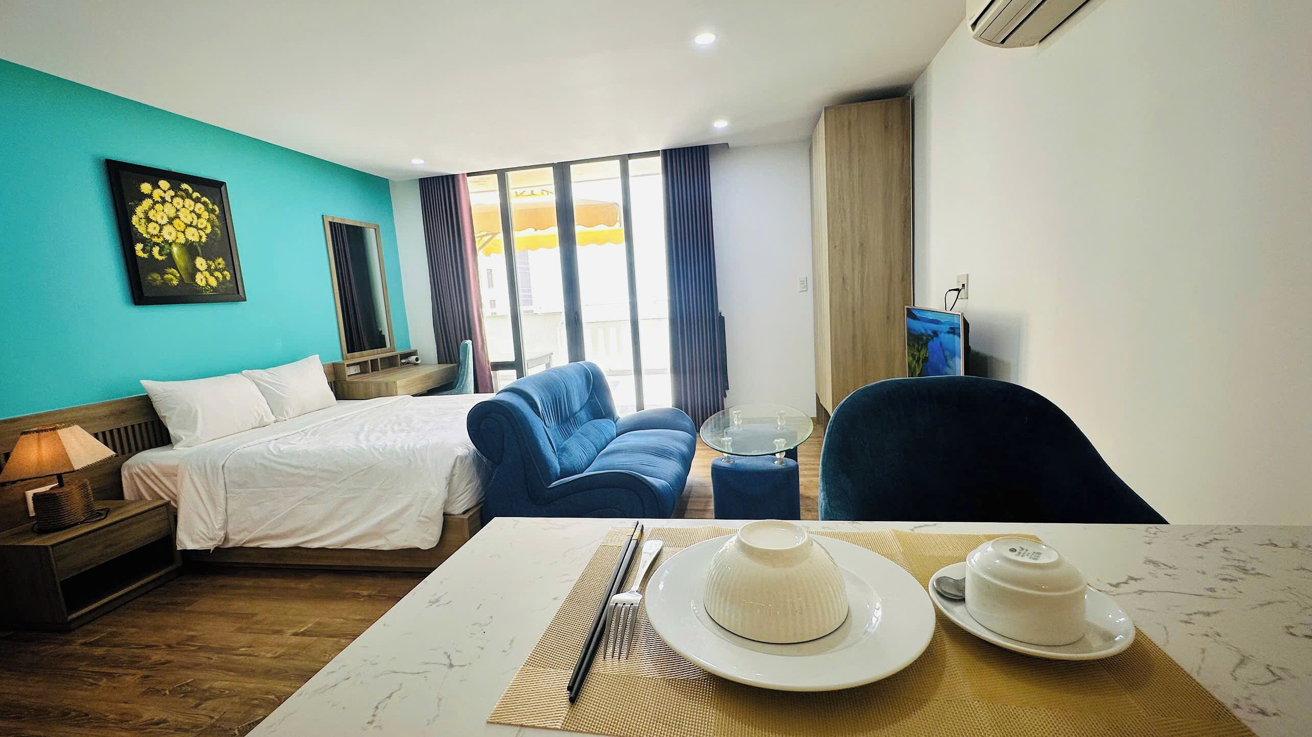 Une chambre d’hôtel avec un lit, un canapé bleu, une table à manger avec une assiette et une tasse, et une grande fenêtre.