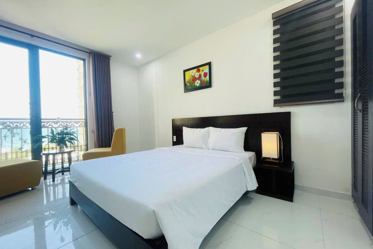 A hotel room with a large bed, a bedside table with a lamp, a chair, a balcony with curtains, and a painting on the wall.