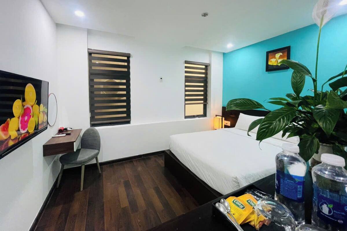 A hotel room with a bed, a desk, a chair, a window with blinds, a wall-mounted TV, a plant, and a tray with bottled water and snacks.