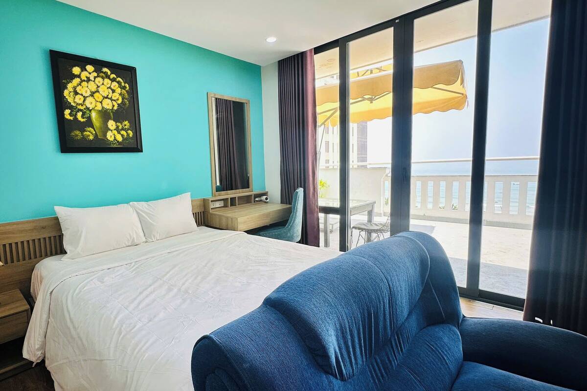 A hotel room with a bed, a desk, a chair, a large window with a view of the sea, and a painting on the wall.